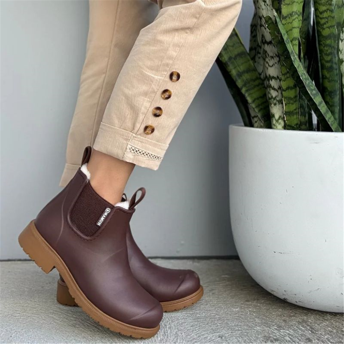 Brown rubber boots worn with beige pants next to a potted plant.